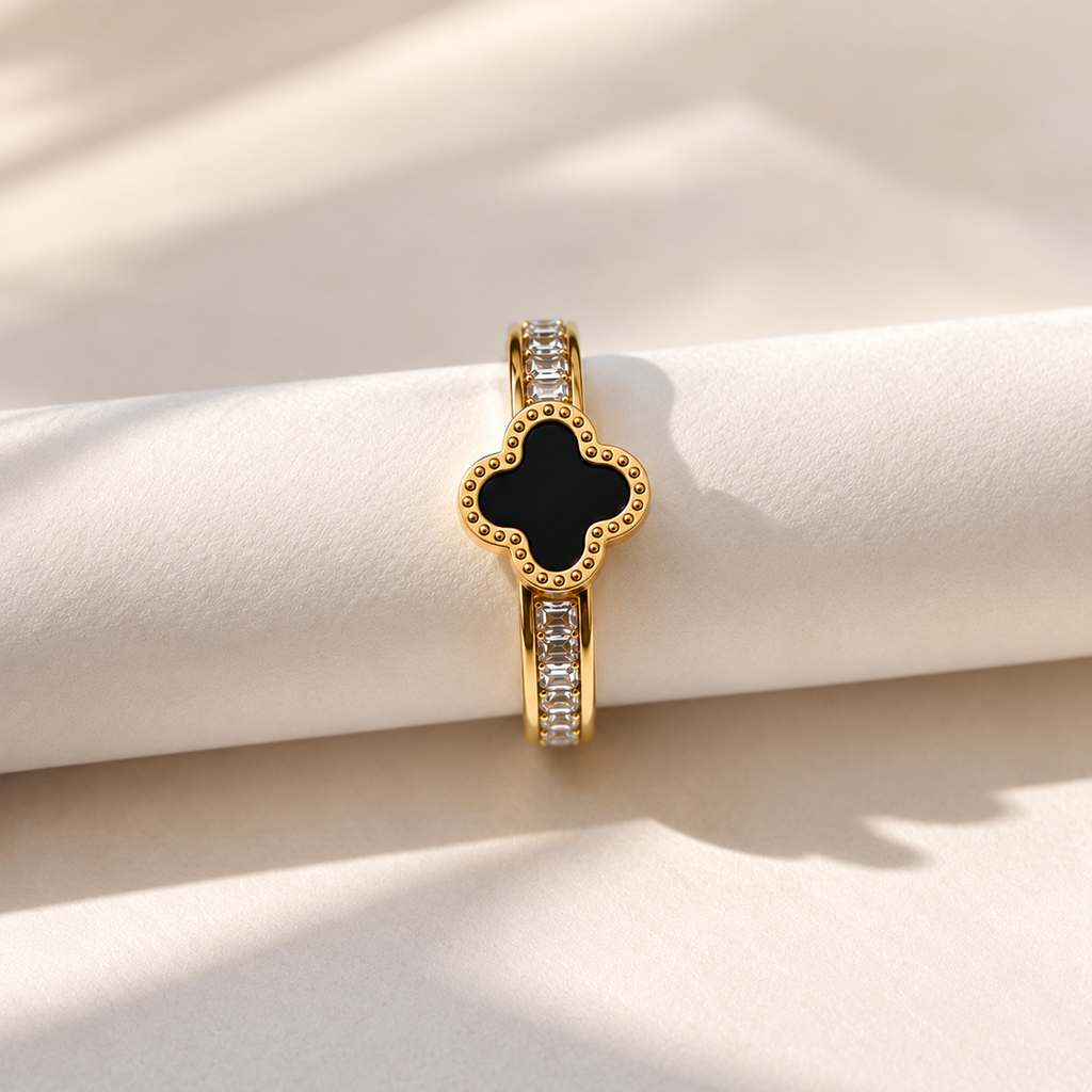 Gold-tone ring with a black four-leaf centre and clear sparkling accents on an ivory surface.