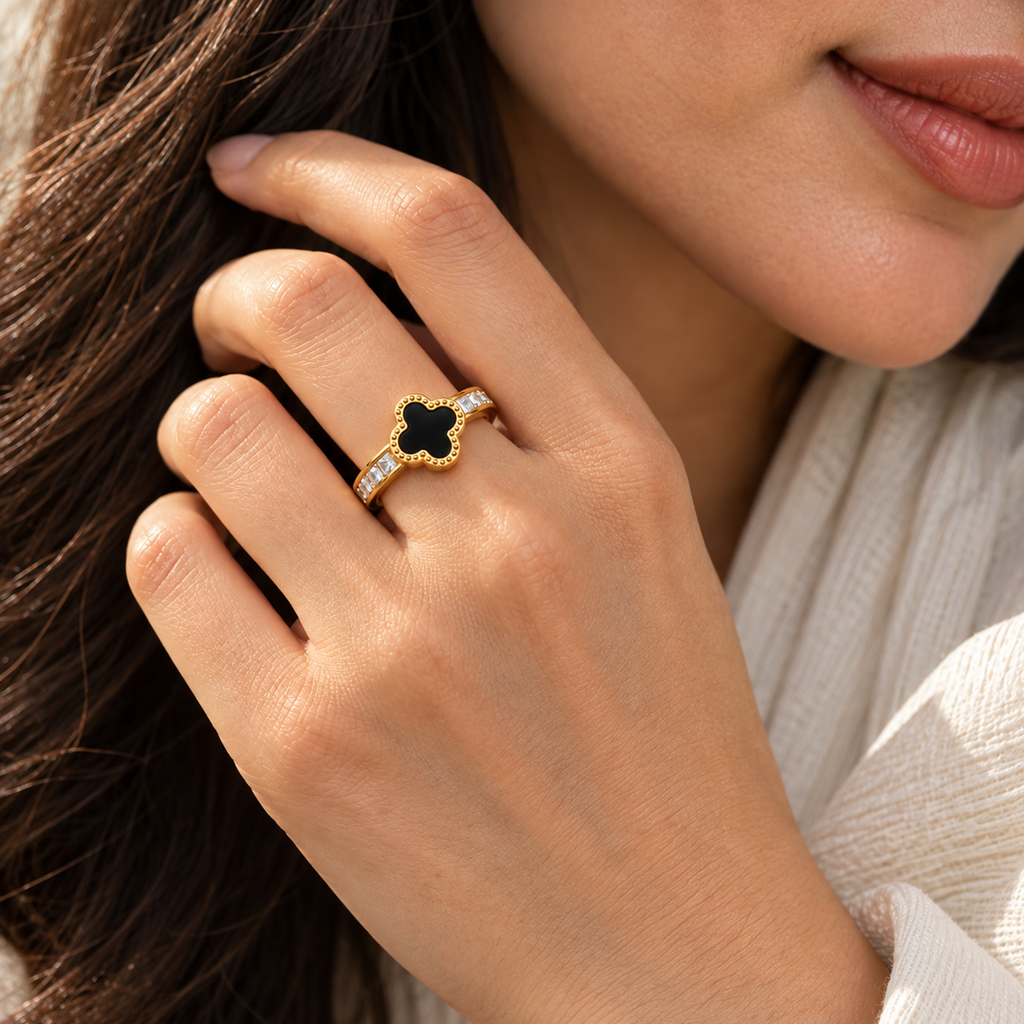 Noir Clover gold-tone ring with a black four-leaf clover center and sparkling clear accents, worn on a woman’s finger in a close-up lifestyle setting.
