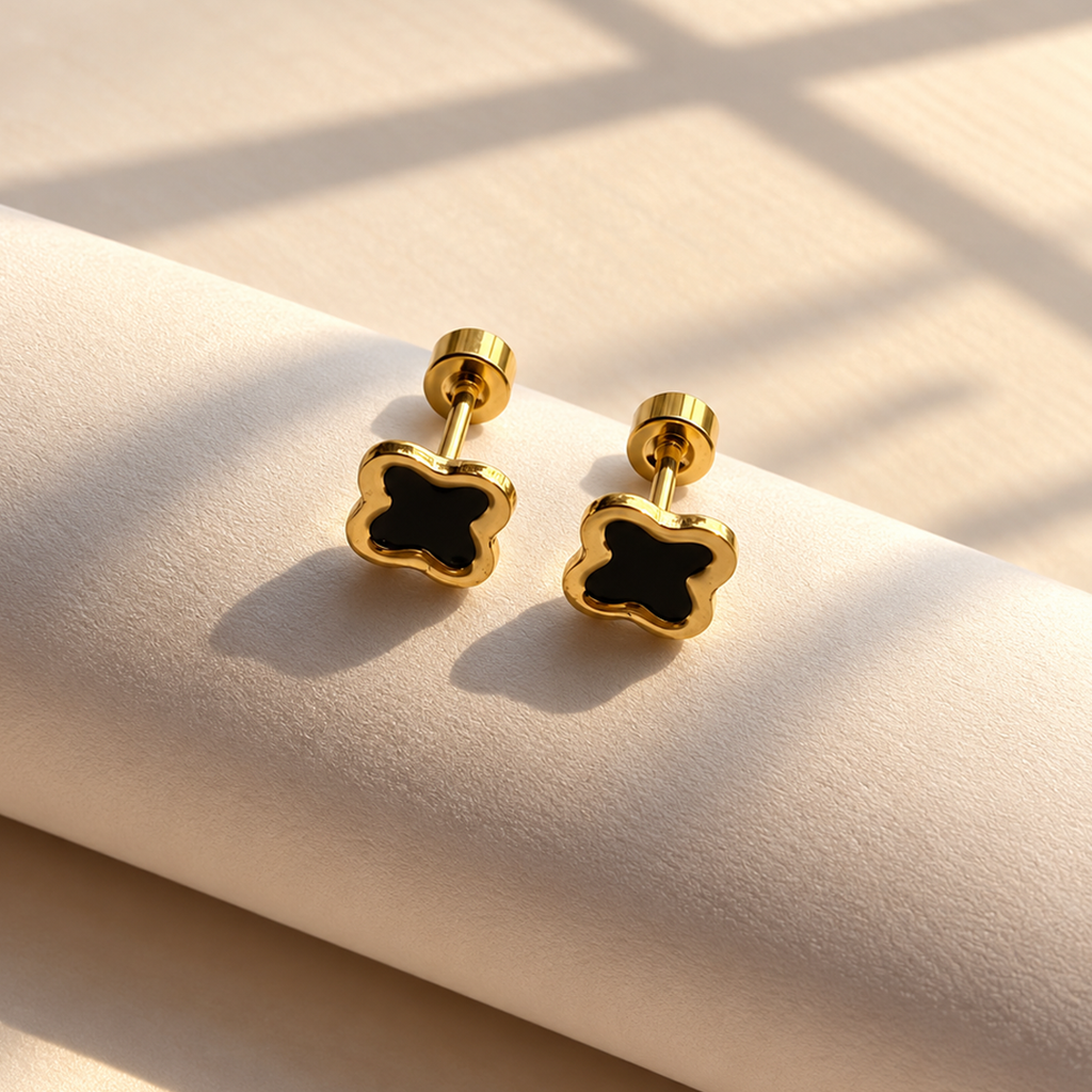 Gold-tone clover stud earrings with black centre details displayed on soft ivory fabric.
