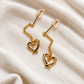 Amour Heart Drop Earrings by Ranaai featuring polished gold-tone heart charms and delicate chain detailing on soft ivory fabric.