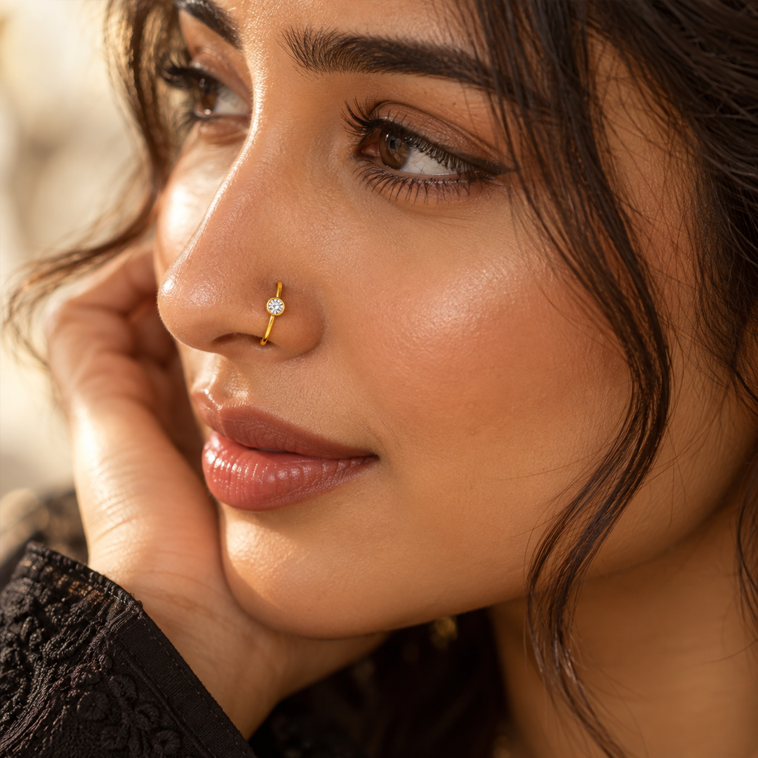 Gold-tone nose ring with a round clear stone worn on the nose, shown in a close-up beauty portrait with warm, soft lighting.